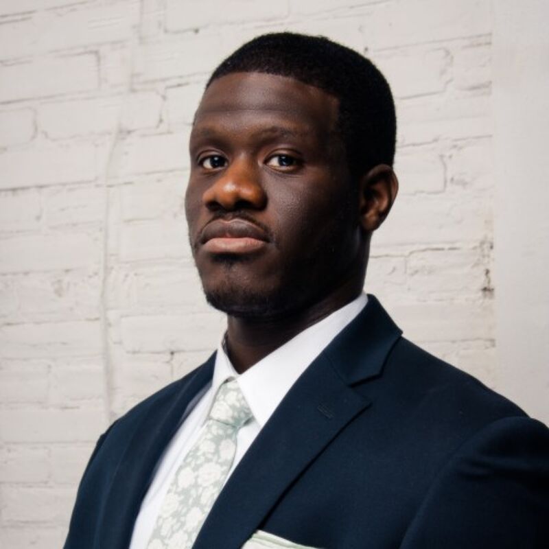 The image shows a man in a suit posing for a portrait. He has short black hair and a serious expression. The man is wearing a dark suit, a light-colored tie with a floral pattern, and a white pocket square. The background is a white brick wall.